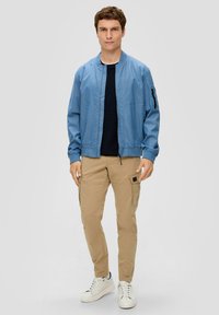 Lichtblauwe bomberjack met een rits, elastische manchetten, gecombineerd met beige cargobroek. Model draagt witte sneakers. Eenvoudig, functioneel ontwerp.