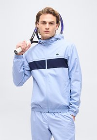 Νέος άνδρας σε ανοιχτό γαλάζιο φόρμα κρατά ρακέτα τένις στον ώμο, στέκεται μπροστά σε απλό λευκό φόντο.