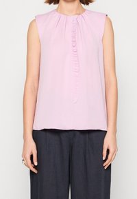 Top sin mangas de color rosa claro con un escote redondeado fruncido y un detalle de volantes verticales en el centro. Tejido suave y liso.
