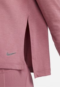 Nahaufnahme eines mauvefarbenen Langarmoberteils mit seitlichem Schlitz und kleinem grauen Nike-Logo oberhalb des Taillenbunds von mauvefarbenen, gerippten Leggings.