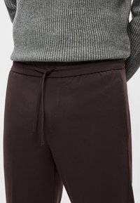 Calças de ganga castanho escuro com cintura elástica e cordão. O tecido parece liso com um leve brilho, combinado com um suéter cinza canelado.