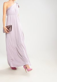 Lila maxiklänning med en axel och elastisk midja, flödande tyg, tillsammans med en texturerad silverclutch och rosa högklackade skor.