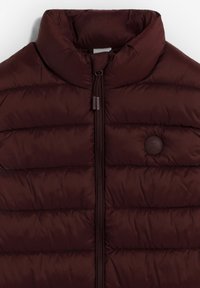 Gilet imbottito bordeaux con colletto alto, cuciture orizzontali, zip frontale e un patch circolare con logo sul lato sinistro. Materiale liscio e strutturato.