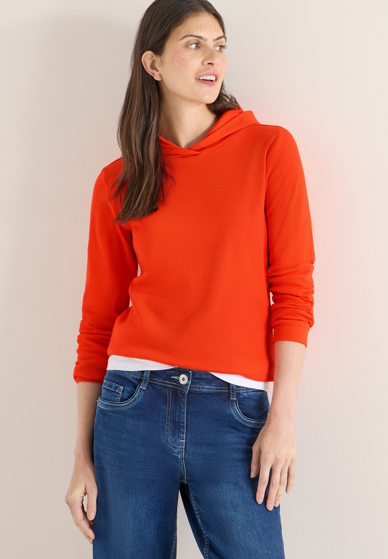 Femme aux longs cheveux bruns portant un sweat à capuche orange vif et un jean bleu, debout contre un mur clair uni, regardant sur le côté.