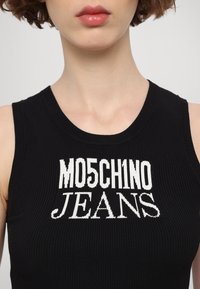 Melnas ribotas krekliņš ar baltu grafikas tekstu "MO5CH1NO JEANS" pāri krūtīm. Kakla izgriezums ir apaļš un pieguļošs.