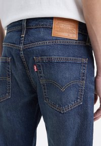 Jeans de mezclilla azul oscuro con costuras visibles, parche de cuero en la cinturilla y dos bolsillos traseros con un diseño curvado característico.
