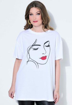 Mujer joven con cabello castaño ondulado, lleva pendientes de aro dorados y una camiseta blanca de gran tamaño con un dibujo minimalista de un rostro con labios rojos.