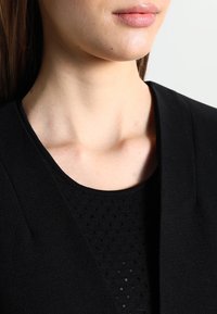 Ženská dolní část obličeje a krku v černém texturovaném topu a černém saku na světlém pozadí.