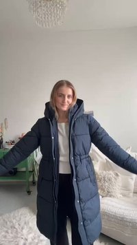 Abrigo acolchado azul oscuro con capucha, con un diseño acolchado, cintura elástica y bolsillos laterales, usado sobre un suéter claro y pantalones negros.