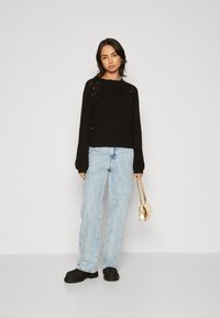 Zwarte gebreide trui met een ronde hals en raglan mouwen, gecombineerd met lichtblauwe baggy jeans en zwarte sneakers. Houdt een kleine gouden handtas vast.