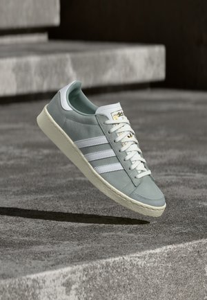 Γκρι σουέτ αθλητικό παπούτσι με λευκές ρίγες, ελαστική σόλα, λευκό κορδόνι και γλώσσα. Διαθέτει χρυσό λογότυπο κοντά στο επάνω μέρος. Πλέει πάνω από μια υφασμάτινη επιφάνεια.