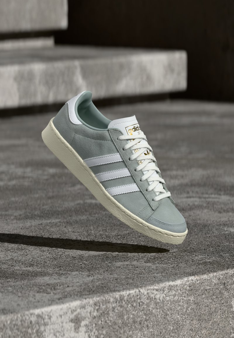 Grijze suède sneaker met witte strepen, rubberen zool, witte veters en tong. Heeft een gouden merklogo vlakbij de bovenkant. Zwevend boven een getextureerd oppervlak.