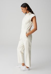 Combinaison en denim blanche à manches courtes, avec boutonnière à l'avant, poche poitrine et poches latérales ; associée à des baskets blanches. Texture lisse.