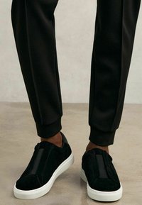Sneakers slip-on in suede nero con pannelli elastici laterali e suole in gomma bianche, abbinate a joggers neri a gamba tapered. Texture liscia, design minimalista.