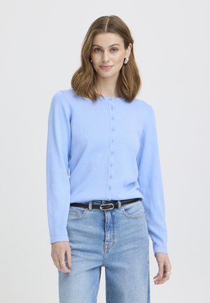 Žena s hnedými vlasmi po ramená má na sebe svetlomodrý zapínaný kardigán, strieborné kruhové náušnice, svetlomodré rifle a čierny opasok na bielom pozadí.