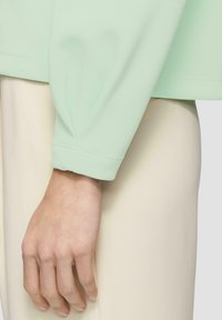 Mintgroene top met een losse pasvorm, puffy mouwen en een gladde textuur, gecombineerd met crèmekleurige broeken. Natuurlijke hand rust dichtbij.