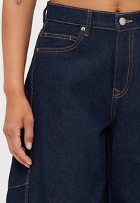 Pantaloni largi din denim albastru închis, cu cusături contrastante, având buzunare frontale și închidere cu buton. Textură netedă și croială lejeră.