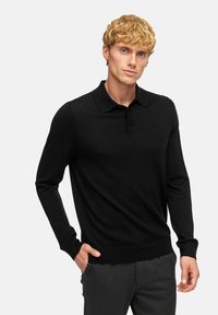 Polo shirt nero a maniche lunghe realizzato in un tessuto morbido, con colletto classico e patta con tre bottoni. Indossato con pantaloni grigio scuro.