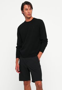 Ung mann i svart langermet genser og svarte shorts som står med den ene hånden i lommen, og ser til venstre mot en hvit bakgrunn.