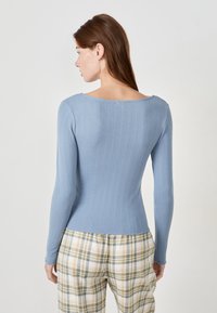 Mujer con cabello castaño que lleva un top de manga larga azul claro de canalé y pantalones a cuadros amarillo-verde, vista desde atrás contra un fondo liso.