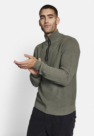 Khakifarbener Strickpullover mit hohem Kragen und halblanger Reißverschlussöffnung. Verfügt über eine gerippte Textur und enganliegende Bündchen, kombiniert mit dunklen Hosen.