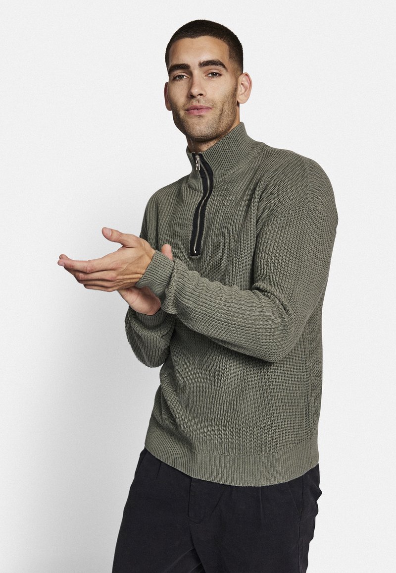 Maglione a maglia verde khaki con colletto alto e chiusura a metà zip. Presenta una texture a costine e polsini aderenti, abbinato a pantaloni scuri.