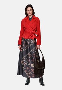 Red suede jacket with a tie waist, patterned long skirt in dark shades, and a dark handbag with fringe accents. Black heeled boots.