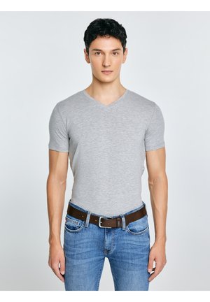 Jauns vīrietis ar īsiem tumšiem matiem, tērpies piegulošā pelēkā T-kreklā ar V veida izgriezumu, zilos džinsos un brūnā ādas jostā, stāv pret vienveidīgu fonu.