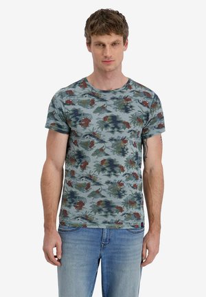 Young man wearing a blue floral-patterned T-shirt and light blue jeans, standing against a plain white background.