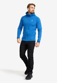 Blå huva jacka med full dragkedja och långa ärmar, tillsammans med svarta cargobyxor som har fickor och dragkedjor. Svarta skor fulländar stilen.