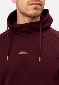 Haut de hoodie bordeaux avec un col montant, affichant le texte jaune "ICONIC EXPERIENCE" et des accessoires orange sur les cordons de serrage. Tissu doux et texturé.