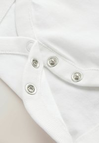 Nærbillede af hvidt stof med tre metaltrykknapper delvist fastgjort på en beklædningskant, der viser syning og tekstur.