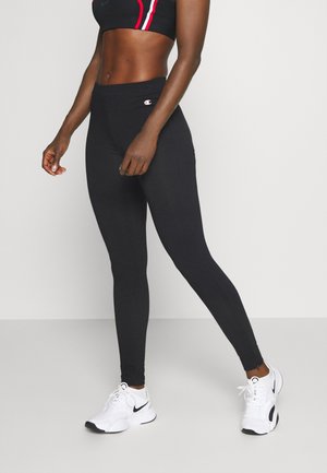 Atletisk person, der bærer sorte leggings, sort sports-bh med røde og hvide striber og hvide Nike-sneakers, står på en ensfarvet baggrund.