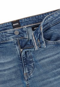 Mörkblå denimjeans med dragkedja och knappstängning, i orange sömmar och en etikett som visar "BOSS" och "DELAV."