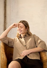 Jeune femme aux yeux fermés et souriante, assise confortablement dans une chaise en bois, portant une chemise beige boutonnée et un short noir.