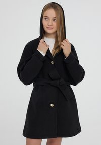 Giovane donna che regola il cappuccio su un cappotto nero con cintura e bottoni, indossando una maglietta bianca con polsini a volant, in piedi su uno sfondo neutro.