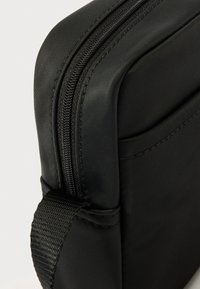 Fekete párnázott tok sima textúrával, erős cipzárral és fonott pánttal. Kompakt kialakítás, alkalmas elektronikai eszközökhöz.