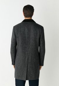 Szürke tweed kabát, fekete hegyes gallérral, hátul egyetlen szellőzőnyílással és testhez álló szabással, gombos ujjakkal és formázott vállakkal.
