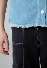 Šviesiai mėlynas džinsinis švarkas su nusidėvėjusiu apvadu, derinamas su juodomis kelnėmis, turinčiomis baltus siūlus ir kišenes. Šonap žiūrint dalinai matoma ranka.