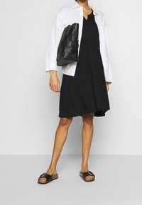 Robe noire à volants avec manches courtes, associée à une chemise blanche boutonnée et un sac tissé noir. Sandales plates noires avec accents métalliques.
