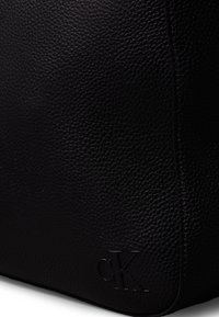 Musta nahkabag, jossa on kivinen pinta, rakenteellinen muoto ja brodeerattu "CK"-logo. Siistit linjat ja vähäiset yksityiskohdat.