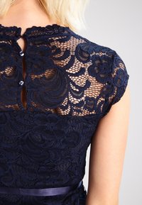 Marinblå spetsklänning med ett blommönster, korta ärmar och en nyckelhål rygg med två knappar. Inkluderar ett midjeband i satin.