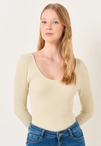 Langärmliger, gerippter Body in hellem Beige, mit einem runden Ausschnitt. Kombiniert mit blauen Jeans. Glatte Textur und taillierte Passform.