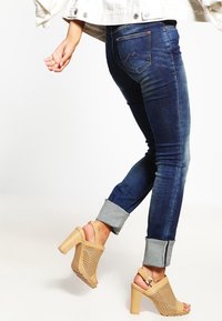 Vaqueros en denim de color azul oscuro con dobladillo, combinados con sandalias de tacón alto color beige, perforadas y con un hebilla en la correa del tobillo.
