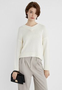 Krémový pletený sveter s v-neck výstrihom a rebrovanými manžetami a lemom, v kombinácii s svetlo béžovými nohavicami so zvýšeným pásom a malou čiernou kabelkou so zlatým uzáverom.