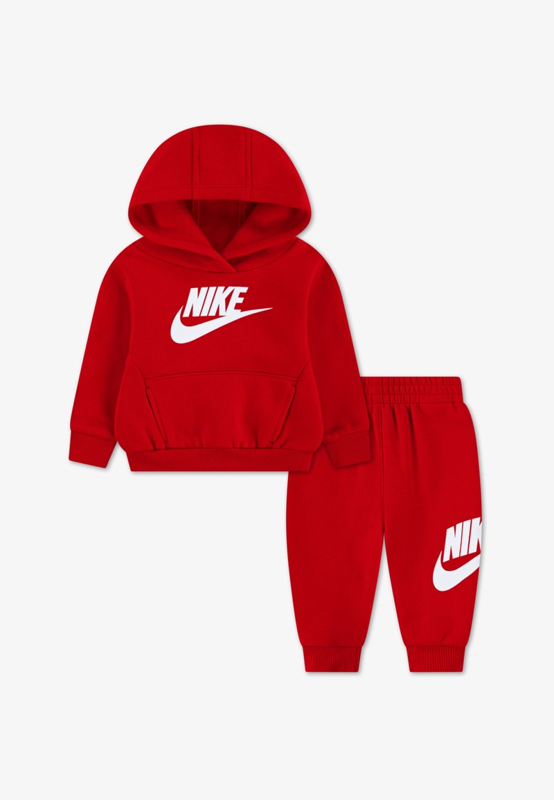 Rdeč Nike trenirka z jopico s kapuco in sprednjim žepom ter ujemajoče se trenirke. Oba kosa imata velik bel Nike logotip.