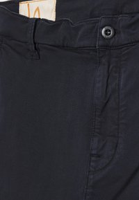 Gros plan sur la taille d'un pantalon bleu marine avec bouton, passant de ceinture et étiquette partielle visible à l'intérieur de la ceinture.