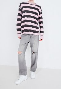 Camicia a maniche lunghe a righe in tessuto rosa e nero, abbinata a jeans strappati grigi chiari e sneakers bianche, su uno sfondo neutro.