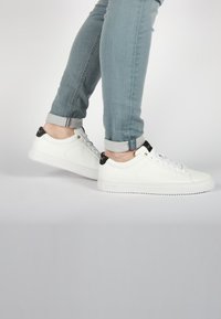 Sneakers bianchi in pelle con punta rotonda, dettagli neri sul tallone e lacci. Indossati con jeans azzurri a risvolto, che mettono in risalto un design elegante e minimale.
