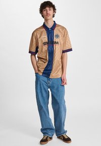 Kortærmet poloshirt i guld og navy med diamantmønster, med et logo og farvede detaljer på kraven og ærmerne.